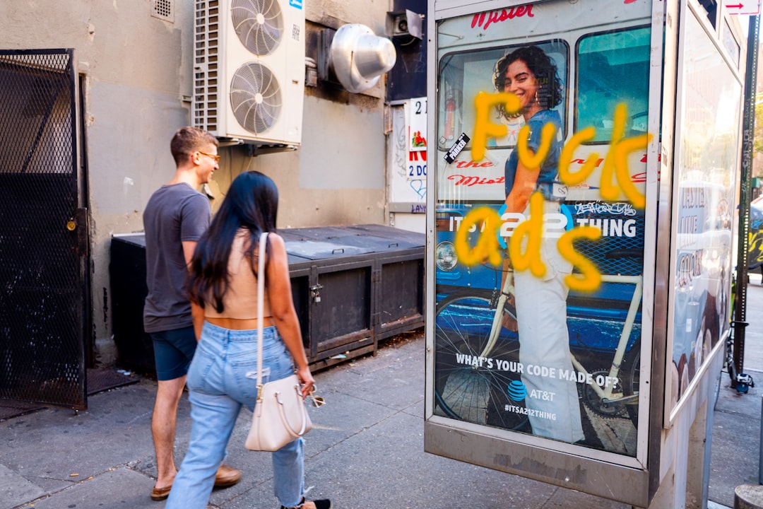 Two pedestrians walk past a bus shelter advertisement that has been tagged with the phrase “Fuck ads” in thick yellow spray paint, partially covering a commercial poster that still shows “WHAT’S YOUR CODE MADE OF?” and AT&T branding, capturing a tense collision between public space, advertising, and rebellion.